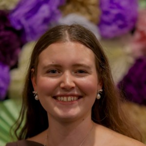 headshot of a young woman with long light brown hair, parted in the side, smiling with out of focus purple and white flowers in the background